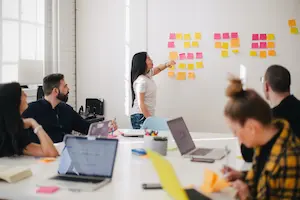 A meeting room with a few people seated. Someone is standing at the whiteboard moving sticky notes around.